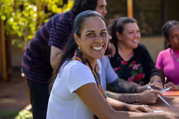 Close-up of aboriginal students and their tutor sitting outdoors in Australia. One of the female students is looking at the camera and smiling.