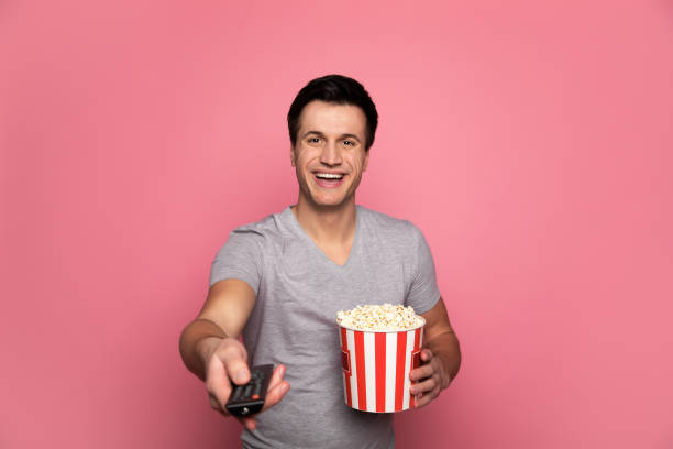 Home cinema. Close-up photo of a happy man in casual outfit, who is holding a remote controller in his right hand and a tube of a popcorn in his left hand and smiling. Home cinema. Close-up photo of a happy man in casual outfit, who is holding a remote controller in his right hand and a tube of a popcorn in his left hand and smiling. popcorn snack bowl isolated stock pictures, royalty-free photos & images