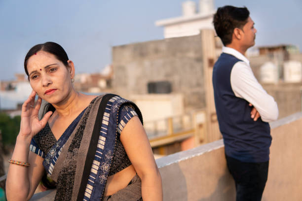 Indian mature couple standing separately after having fight. Indian mature woman looking upset, despondent after having fight with her husband standing in background on rooftop at day time. husband and wife stock pictures, royalty-free photos & images