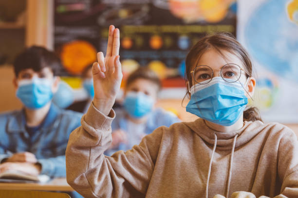 High school students at school, wearing N95 Face masks. High school students at school, wearing N95 Face masks. Sitting in a classroom and listening to teacher. Teen girl is rising hand. teenage high school girl raising hand during class stock pictures, royalty-free photos & images
