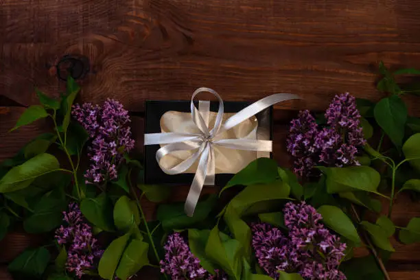 Lilac flowers, black gift box with silver ribbon on wooden background flat lay. Text place 8 March Happy Women's, Mother's Day. Purple flower bouquet, greeting card copy space,website banner top view.