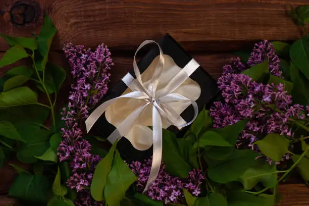 Lilac flowers, black gift box with silver ribbon on wooden background flat lay. Text place 8 March Happy Women's, Mother's Day. Purple flower bouquet, greeting card copy space,website banner top view.