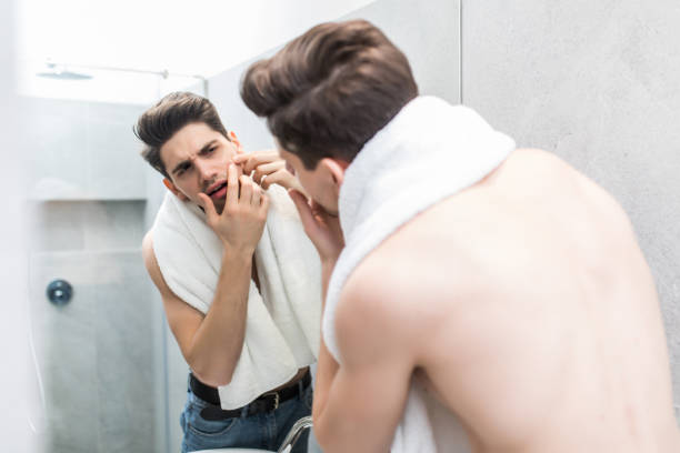 istock Young man standing in front of mirror and touching his face in bathroom 1225031024