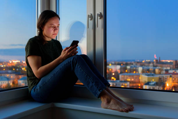 istock Young Woman using phones sitting on windowsill at night 1225031516