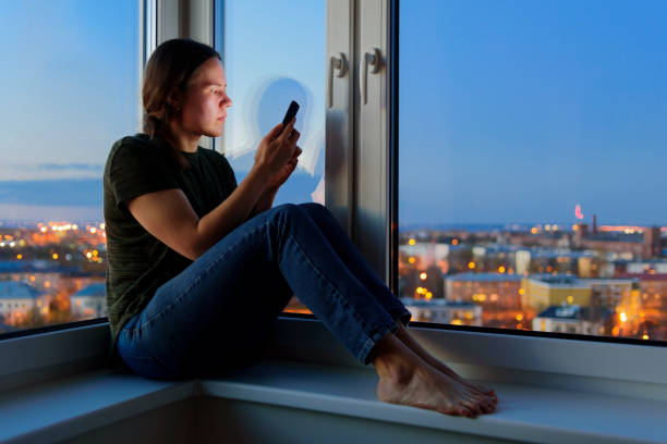 istock Young Woman using phones sitting on windowsill at night 1225031663