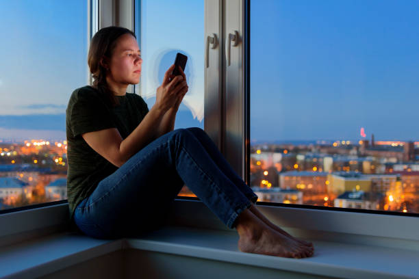 istock Young Woman using phones sitting on windowsill at night 1225031739