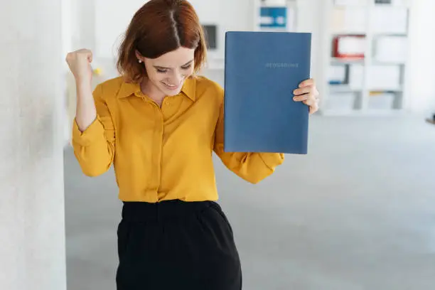 Exultant young woman celebrating getting a job after a successful interview, smiling and punching the air with her fists while holding her CV