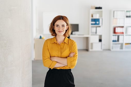 Happy relaxed confident young businesswoman standing with folded arms in a spacious office looking at the camera with a warm friendly smile