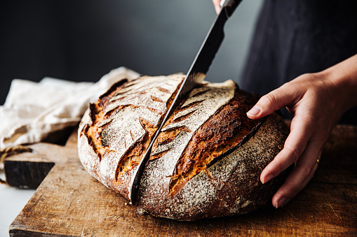 Close-up of woman cutting sourdough bread on board. Midsection of female is with knife and baked bread. She is in kitchen.