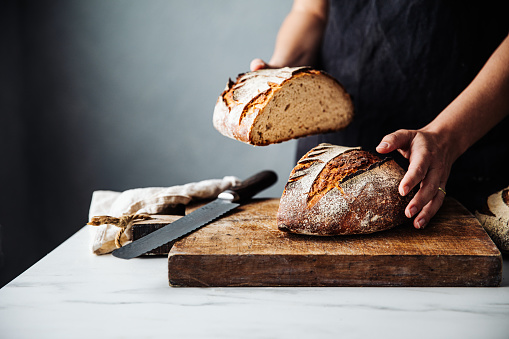 Midsection of woman holding bread with knife on cutting board. Close-up of fresh baked sourdough bread at counter. She is in kitchen.