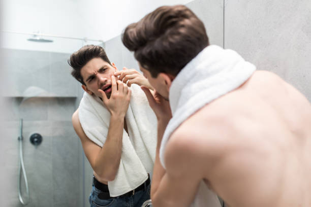 istock Young man standing in front of mirror and touching his face in bathroom 1225040001