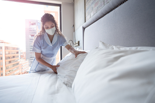 Latin American maid working at a hotel and doing the bed wearing a facemask - COVID-19 pandemic lifestyle concepts
