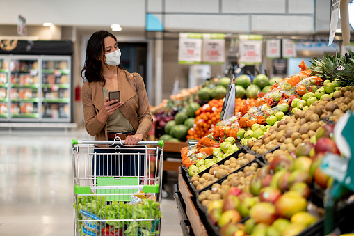 Woman shopping at the grocery store wearing a facemask to avoid the coronavirus while following a list on her cell phone - COVID-19 lifestyle concepts