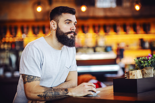 Young bearded tattooed hipster sitting in cafe, holding cup of coffee, thinking and daydreaming.