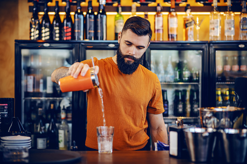 Young attractive muscular Caucasian bearded tattooed barman standing near bar counter and making a cocktail.