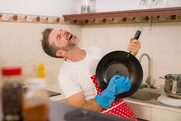 enjoying domestic chores - young cool and attractive man in red apron and washing glove singing song and dancing crazy happy holding pan as if playing guitar excited and carefree stock photo