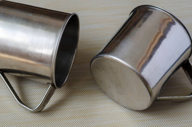 Close-up stainless steel mugs on a textured background. A pair of mugs for hot drinks and food. Dishes for tourism, fishing and hunting. Top view at an angle. Close-up. Selective focus.
