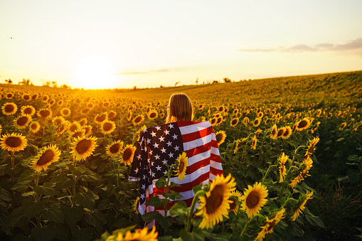 Beautiful girl with the American flag in a sunflower field.  Fourth of July. Sunset light. Patriotic holiday.