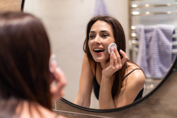 istock Beautiful young woman is cleaning her face using a cotton disc and smiling while looking in the mirror in the bathroom 1225055233