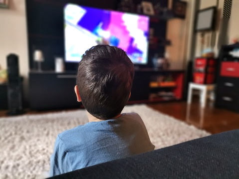 Back view of kid watching TV while relaxing in the living room.