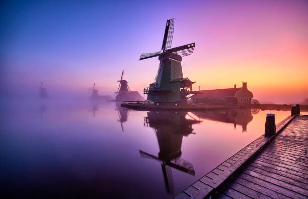 Traditional Dutch windmills in the mist during sunrise along a canal at the Zaanse Schans in The Netherlands. Traditional Dutch windmills in the mist during sunrise along a canal at the Zaanse Schans in The Netherlands. Zaanse Schans is a neighborhood of Zaandam, near Zaandijk, Netherlands. It has a collection of well-preserved historic windmills and houses. The Zaanse Schans is one of the popular tourist attractions of the Netherlands. netherlands windmill stock pictures, royalty-free photos & images