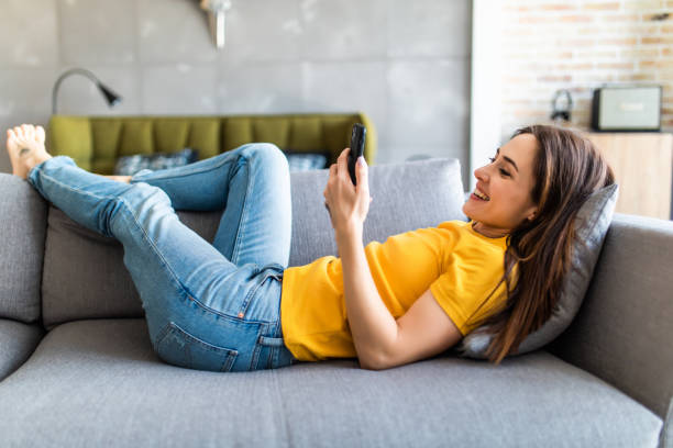 istock Smiling beautiful girl using mobile phone relaxing on a couch at home 1225069411