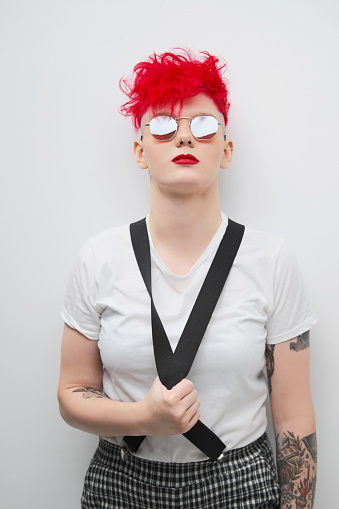 Portrait of a cool fashionable modern young girl. A short haircut with shaved temple. Dyed bright red hair. Red lipstick. Studio photo on a white background. Suspenders on checkered pants and sunglasses.