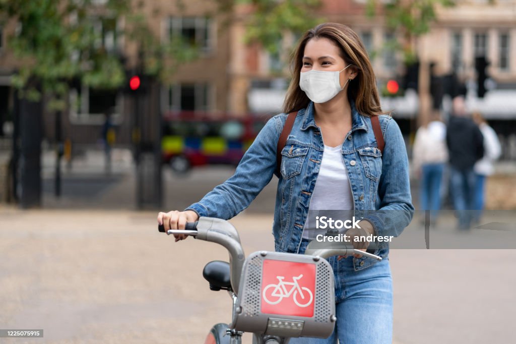 Woman outdoors wearing a facemask and renting a bike Portrait of a happy woman outdoors wearing a facemask to avoid the spread of coronavirus and renting a bike in London â pandemic lifestyle concepts Cycling Stock Photo