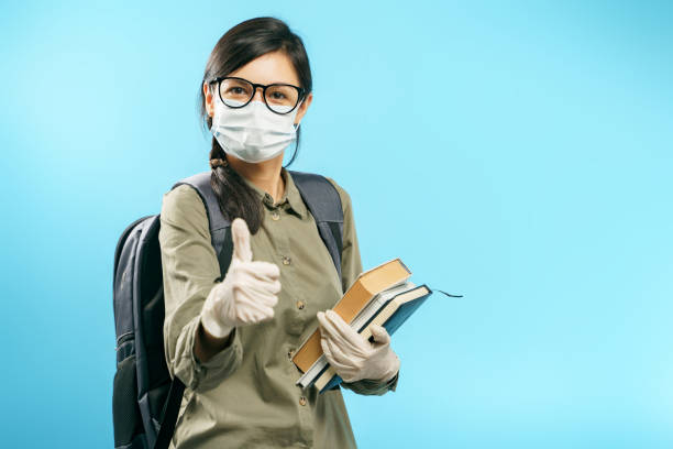 portrait of a female student in a medical protective mask and gloves holding books and showing a thumbs up on a blue background - medical student healthcare and medicine book education imagens e fotografias de stock