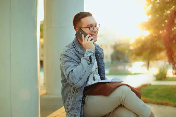 Photo of Young student studying for term exam in the park outdoors. Stylish man talking on cell phone on campus.