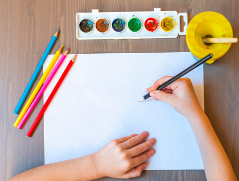 The child's hands are painted with colored pencils on a white sheet of paper on a wooden table. the view from the top. Empty space for your text.