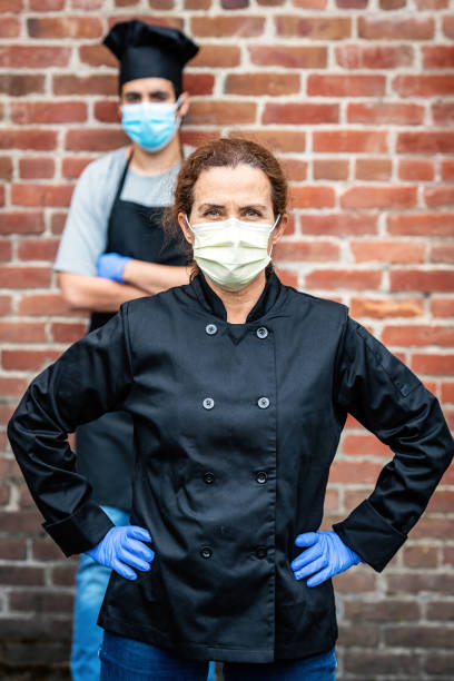 istock Mature female restaurant worker posing with a Young chef coworker wearing both a protective masks and gloves during a break looking at the camera 1225089215