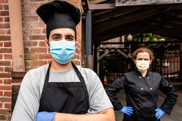 istock Young chef posing with female coworker wearing both a protective masks and gloves during a break looking at the camera 1225091370