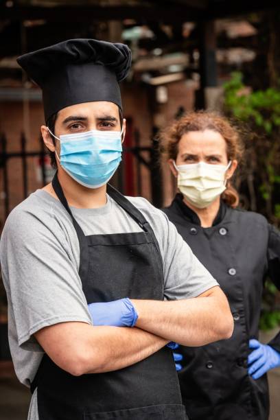 Young chef posing with female coworker wearing both a protective masks and gloves during a break looking at the camera