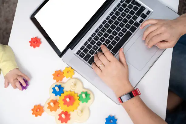 Close up high angle view of hands of mother using laptop for working at home while daughter working with multicolored toy gears. Shot with a full frame mirrorless camera.