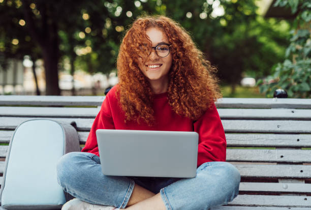 istock Happy red haired student with laptop sitting on bench 1225115746