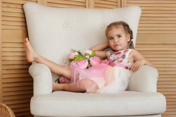 Portrait of pretty little girl in beautiful dress sitting with freedom on the beige armchair with bouquet of pink roses, beauty and love concept, valentine and mother day