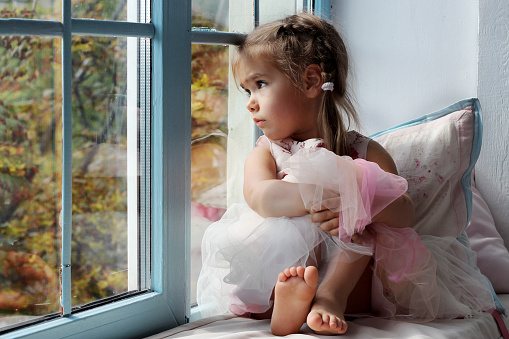 Sad little girl sitting alone at home near the window and looking on the autumn street, indoor portrait, childhood and family concept