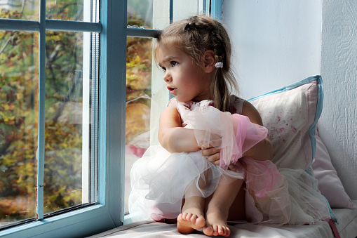 Sad little girl sitting alone at home near the window and looking on the autumn street, indoor portrait, childhood and family concept