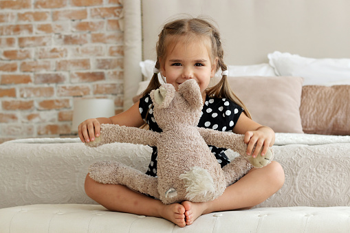 Cute little girl in black and white dress hugging her toy hare on the bed, happy childhood concept, indoor horizontal portrait