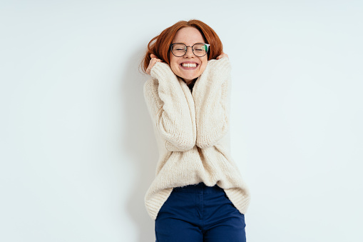 Cute young woman cuddling down into a warm cream colored woollen sweater with a happy smile of pleasure and closed eyes over a white interior wall with copy space