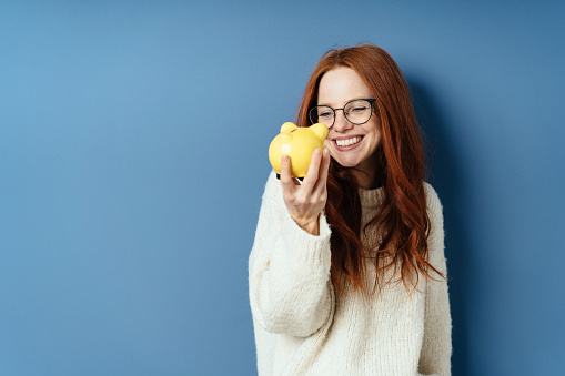 Happy successful young woman smiling at a piggy bank with a look of gleeful anticipation as she imagines what she will be able to do or achieve with her savings on a blue studio background with copy space