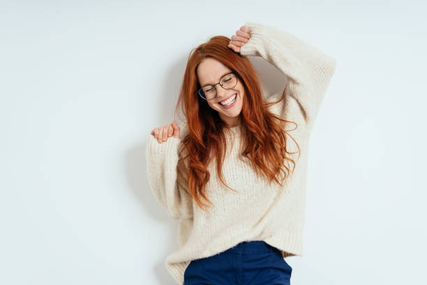 Merry young woman celebrating and dancing Merry carefree young woman wearing glasses and a woollen sweater celebrating and dancing laughing with closed eyes against a white interior wall with copy space one person stock pictures, royalty-free photos & images