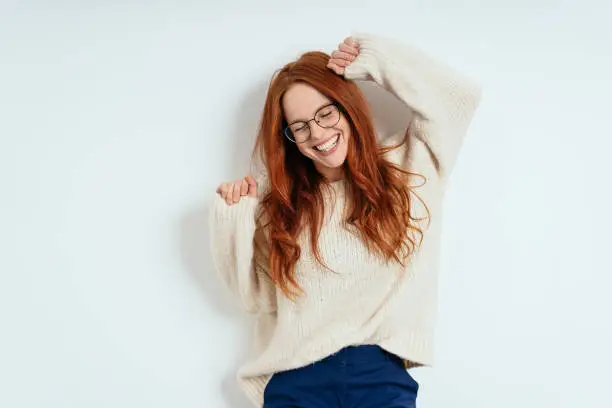 Merry carefree young woman wearing glasses and a woollen sweater celebrating and dancing laughing with closed eyes against a white interior wall with copy space