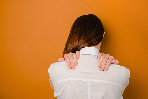 Rear close up view at young brunette woman in white shirt isolated on brown studio background massage back suffering from muscle spasm, unhealthy girl touch neck having backache or sudden strain