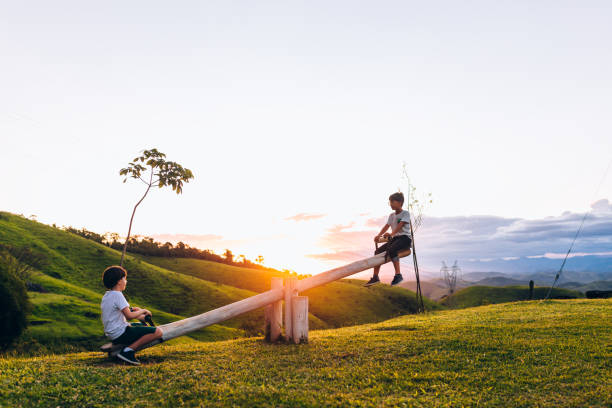 два ребенка, играя на качелях на открытом воздухе во время заката - seesaw стоковые фото и изображения