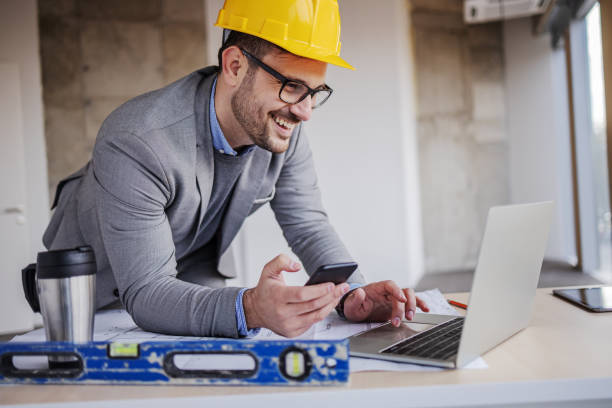 Young smiling architect in suit with helmet on head holding smart phone and using laptop while leaning on table in building in construction process. Young smiling architect in suit with helmet on head holding smart phone and using laptop while leaning on table in building in construction process. architectural model photos stock pictures, royalty-free photos & images