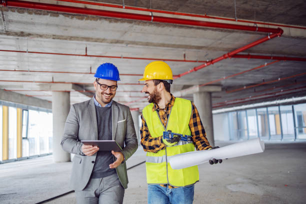 smiling architect in suit with helmet on head talking to contractor while standing in building in construction process. architect holding tablet. - corporate business manager manual worker architect imagens e fotografias de stock