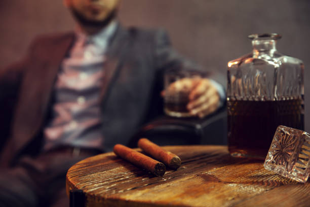 two cigars and a carafe of whisky on wooden table and an elegant man with the glass of whisky in the background - cigar whisky bar cognac imagens e fotografias de stock