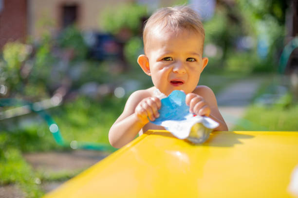 baby holds and eating fruit puree in pouch and looking into the camera with expression in front of the yellow table. on the background is  a green garden on a sunny day in blur. close up stock photo